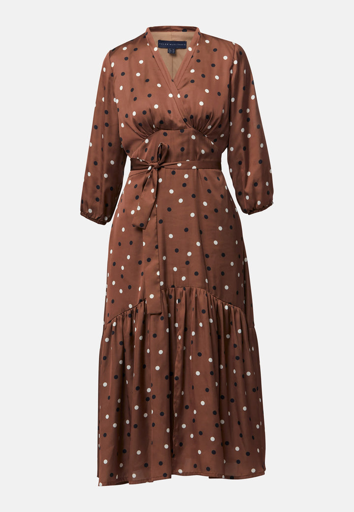 The Beverly Dress in a brown base, black and white polkadot print. This midi dress showcases a flattering fit-and-flare silhouette with a crossover V-neckline and a soft belt to accentuate the waist. The voluminous sleeves add a stylish touch, making it an ideal choice for occasions both casual and dressy.