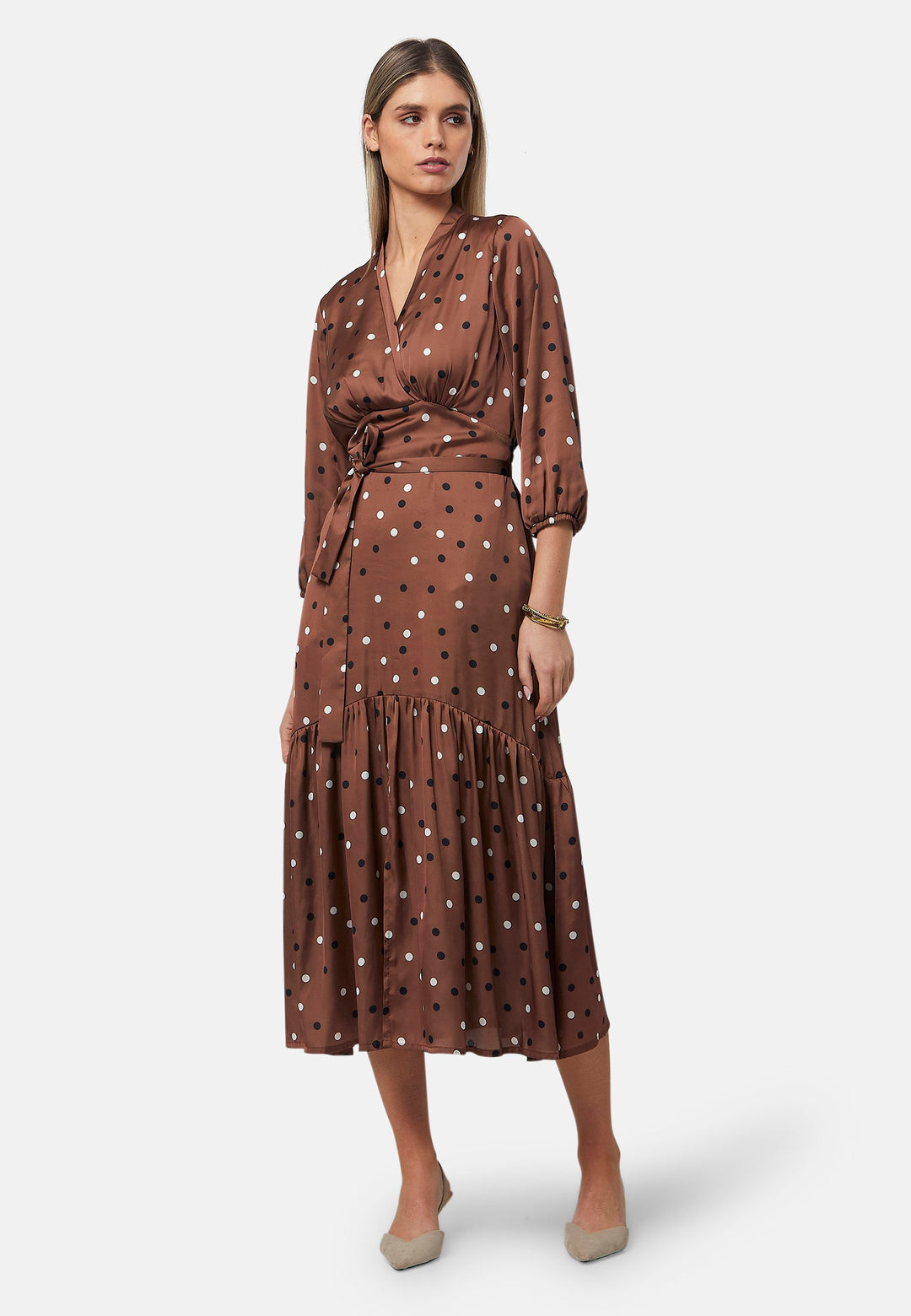 The Beverly Dress in a brown base, black and white polkadot print. This midi dress showcases a flattering fit-and-flare silhouette with a crossover V-neckline and a soft belt to accentuate the waist. The voluminous sleeves add a stylish touch, making it an ideal choice for occasions both casual and dressy.