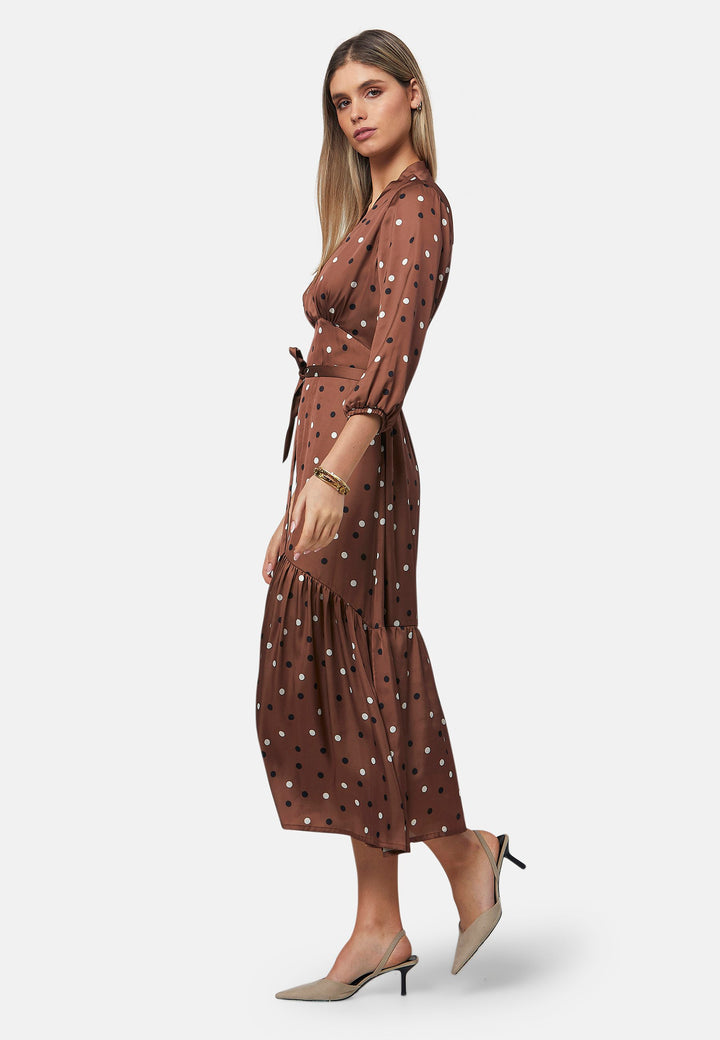 The Beverly Dress in a brown base, black and white polkadot print. This midi dress showcases a flattering fit-and-flare silhouette with a crossover V-neckline and a soft belt to accentuate the waist. The voluminous sleeves add a stylish touch, making it an ideal choice for occasions both casual and dressy.