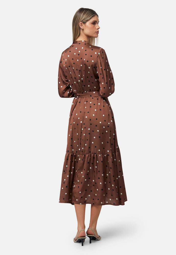 The Beverly Dress in a brown base, black and white polkadot print. This midi dress showcases a flattering fit-and-flare silhouette with a crossover V-neckline and a soft belt to accentuate the waist. The voluminous sleeves add a stylish touch, making it an ideal choice for occasions both casual and dressy.