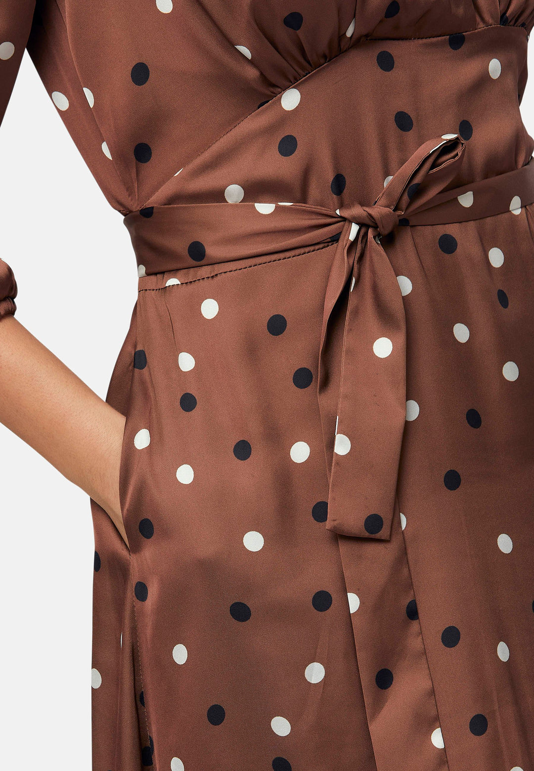 The Beverly Dress in a brown base, black and white polkadot print. This midi dress showcases a flattering fit-and-flare silhouette with a crossover V-neckline and a soft belt to accentuate the waist. The voluminous sleeves add a stylish touch, making it an ideal choice for occasions both casual and dressy.