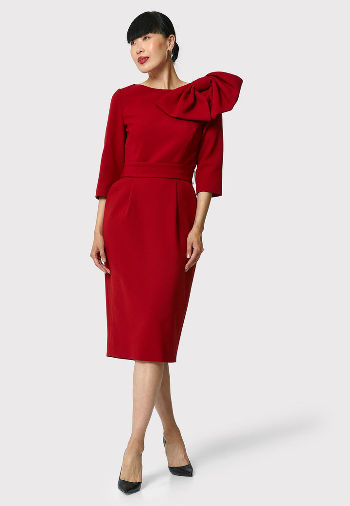 The Isadora Dress in garnet red, a body-skimming silhouette, featuring a slash neckline with a dramatic bow on the shoulder. The dress falls to the mid-calf and back vent ensure ease of movement. Crafted from a fabric blend with a hint of stretch, it offers a comfortable and flattering fit. Ideal for evening cocktails, a night at the theatre, or a stylish winter wedding.