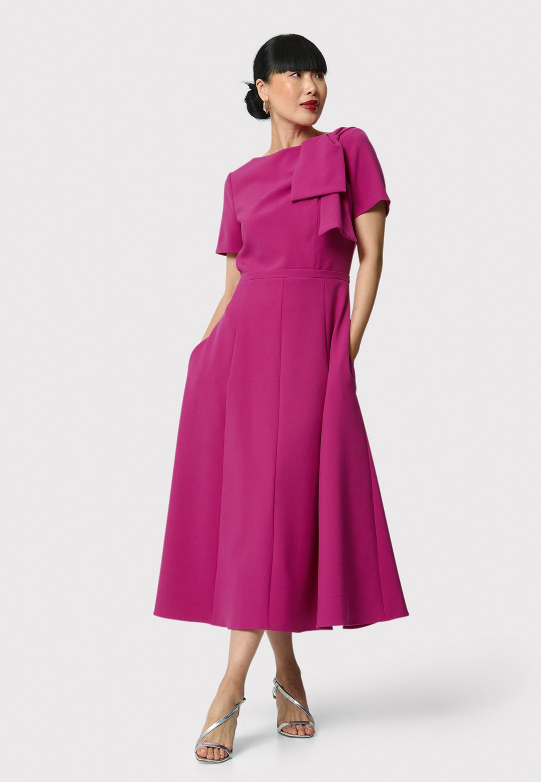 Introducing the Laoise pink dress, a refined statement piece for special occasions. Featuring short sleeves, a fitted bodice, and an elegant shoulder bow detail, this dress has a flattering A-line shape with a cascading paneled skirt. Designed with side seam pockets and a back zip for ease, it combines comfort and sophistication, making it perfect for races, weddings and formal events.