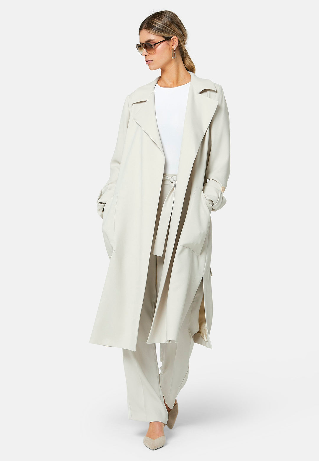 Lydiah, stone trench coat in a fluid satin back crepe. This coat features a relaxed, flowing silhouette, a self-tie belt, and front pockets. The design includes buttoned cuffs and a wide lapel. Pair with coordinating trouser for a chic monochromatic look.