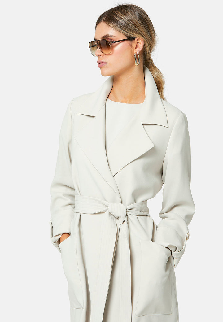 Lydiah, stone trench coat in a fluid satin back crepe. This coat features a relaxed, flowing silhouette, a self-tie belt, and front pockets. The design includes buttoned cuffs and a wide lapel. Pair with coordinating trouser for a chic monochromatic look.