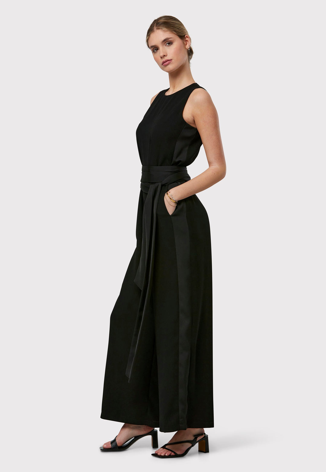 Marie-Claire, a sleeveless black jumpsuit in fluid satin back crepe. Featuring a wide-leg silhouette and satin stripes on the side seams. It comes with a detachable self-fabric belt that can be used to cinch the waist or worn around the neck. Includes practical side pockets for added convenience. Perfect for relaxed yet sophisticated evening dressing.